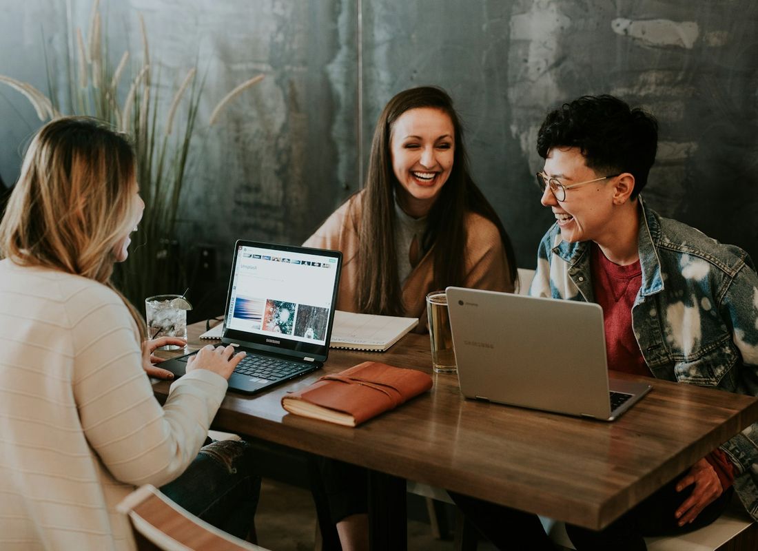 Three planners laughing around a wooden table with two laptops and a notebook